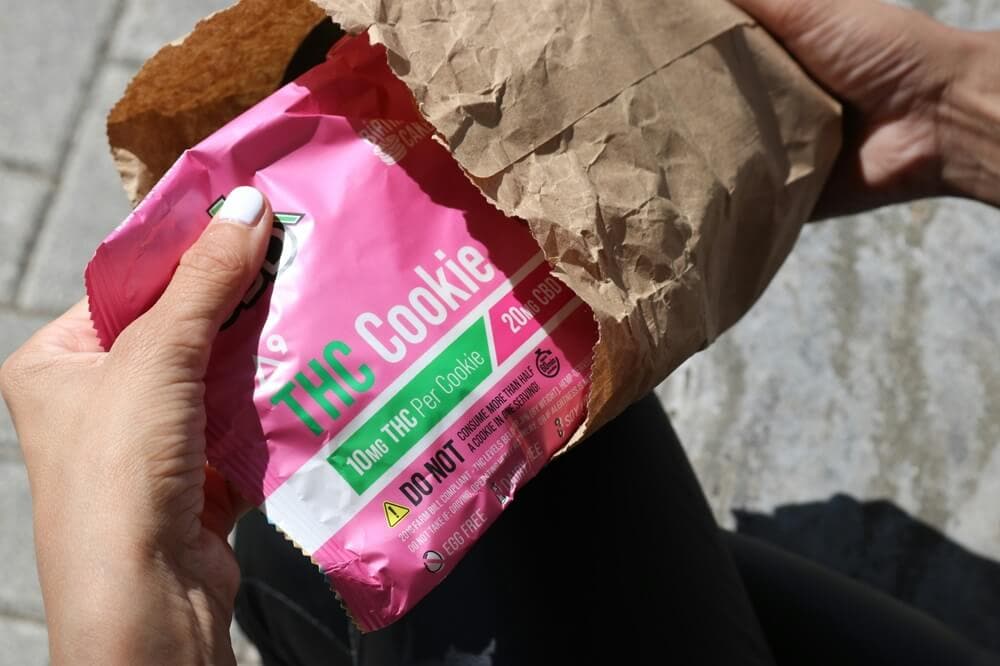 Hand holding pink THC cookie edible pouch emerging from brown paper bag