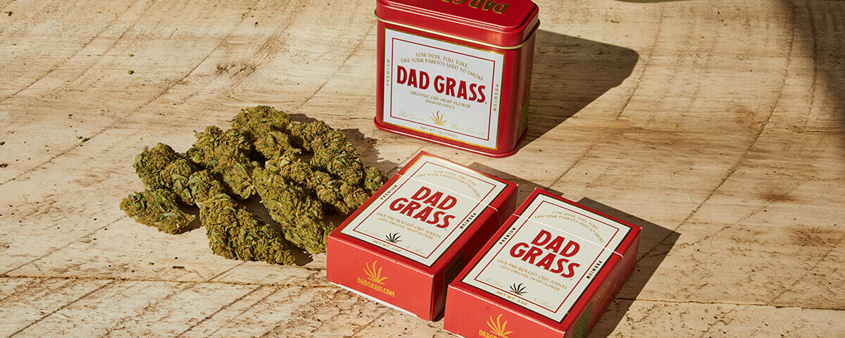 Dad Grass cannabis opaque packaging featuring red boxes and tin container on wood surface.