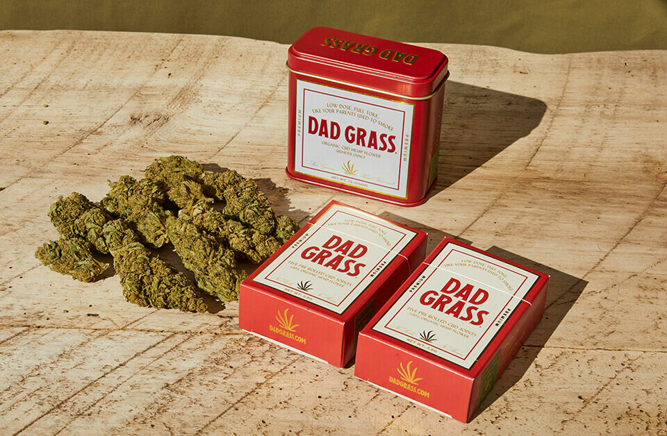 Dad Grass cannabis opaque packaging featuring red boxes and tin container on wood surface.
