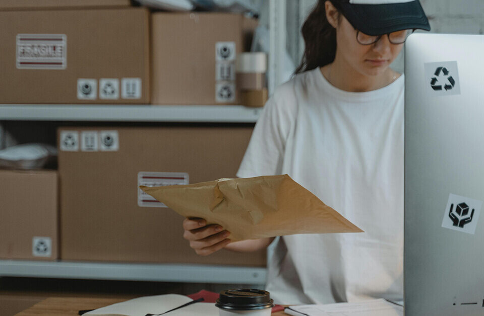 packaging procurement worker packing an order with a brown envelope in front of a computer and cardboard boxes in the background.