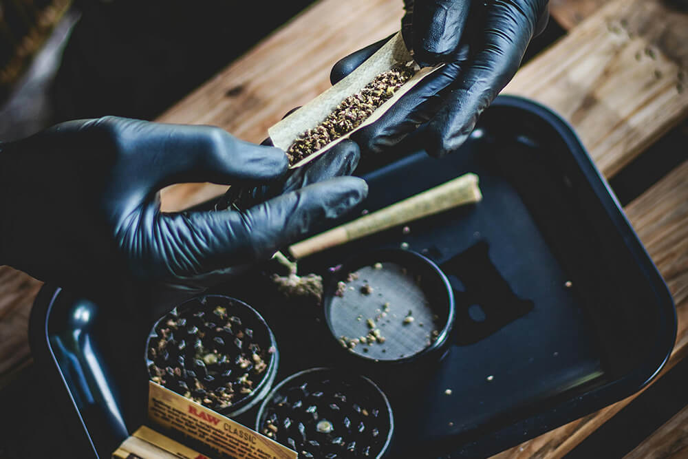 Close-up of gloved hands rolling a joint using a black weed rolling tray with grinders.
