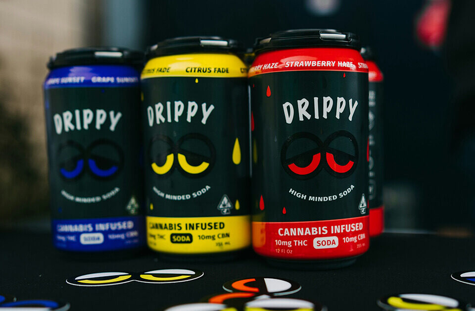 Three cans of Drippy cannabis beverage soda in grape, citrus, and strawberry flavors.