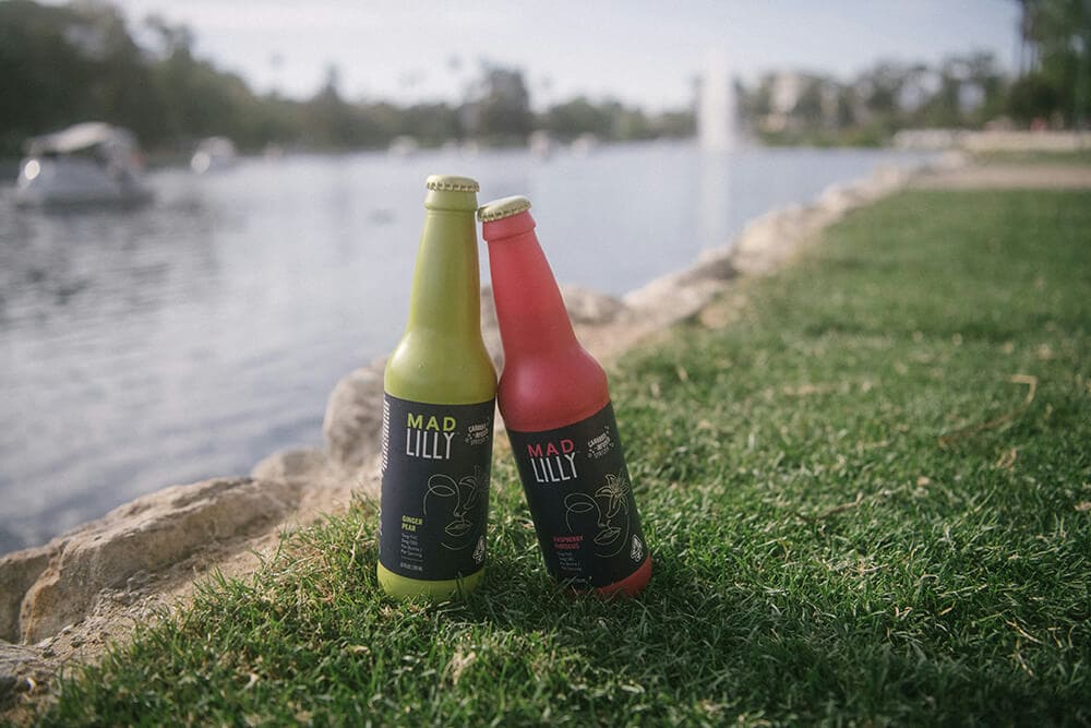 Two Mad Lilly cannabis-infused sparkling beverages in green and red glass bottles by a lakeside.