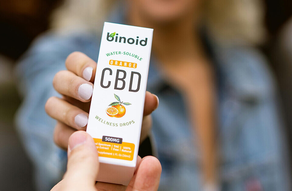 Hand holding Binoid orange CBD wellness drops box, water-soluble supplement medical cannabis packaging.