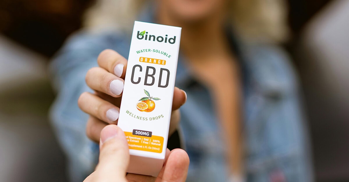 Hand holding Binoid orange CBD wellness drops box, water-soluble supplement medical cannabis packaging.