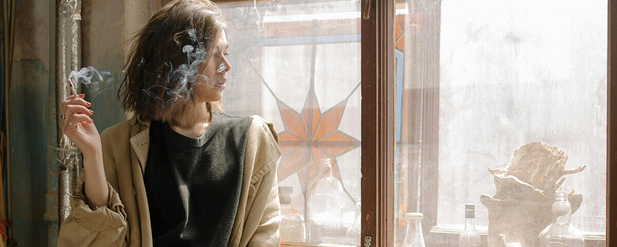 Woman enjoying a mini blunt by the window with sunlight streaming in.