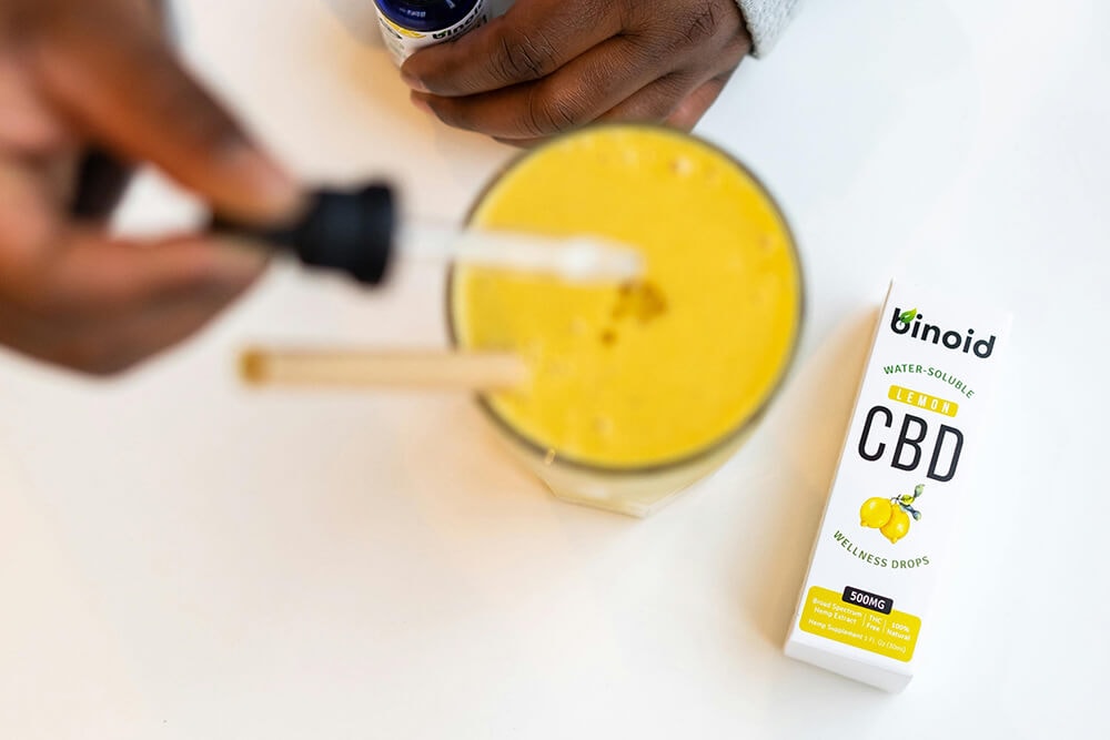 CBD wellness drops packaging placed next to a yellow drink and dropper.