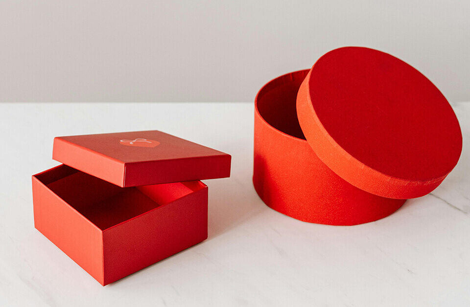 A red square gift box with lid and a round red gift box with lid, both displayed on a marble surface.