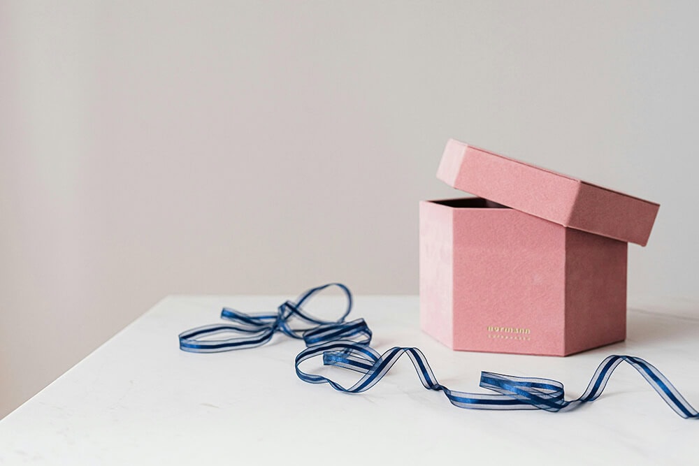 A pink gift box with lid, partially opened and accompanied by a blue ribbon, showing a minimalist, elegant packaging design.