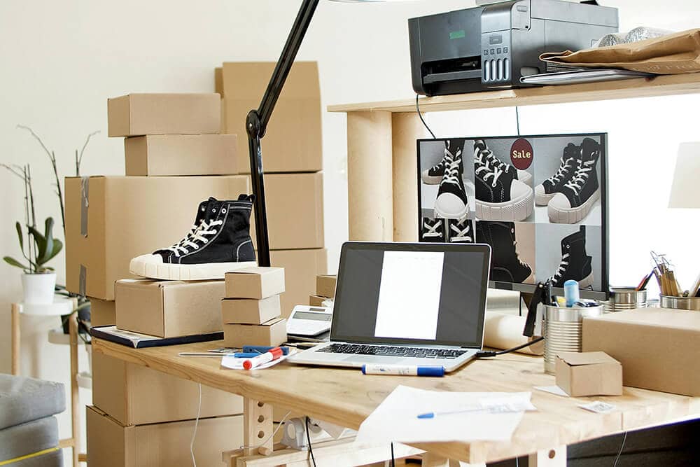 Workspace with stacked e-commerce packaging boxes and sneaker product photography on the desk.