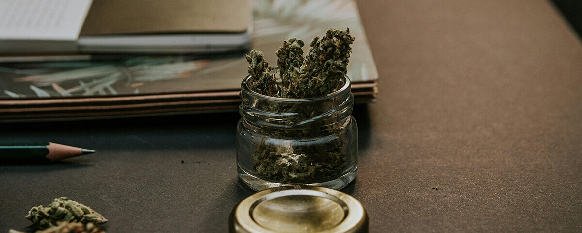 Glass jar used as THC wax packaging placed on a desk among cannabis buds and a notebook.
