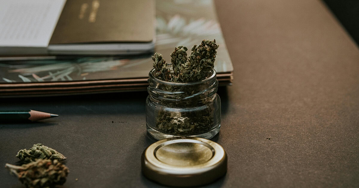 Glass jar used as THC wax packaging placed on a desk among cannabis buds and a notebook.