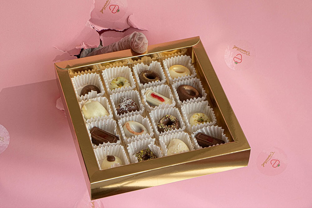 Gold metallic custom chocolate box with a large clear window displaying sixteen assorted pralines on a pink background.