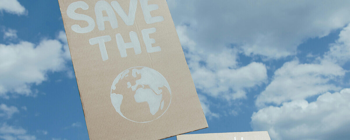 A person holding a cardboard sign that says "SAVE THE EARTH" with an illustration of the globe against a blue sky, representing the environmental goals of sustainable cannabis packaging.
