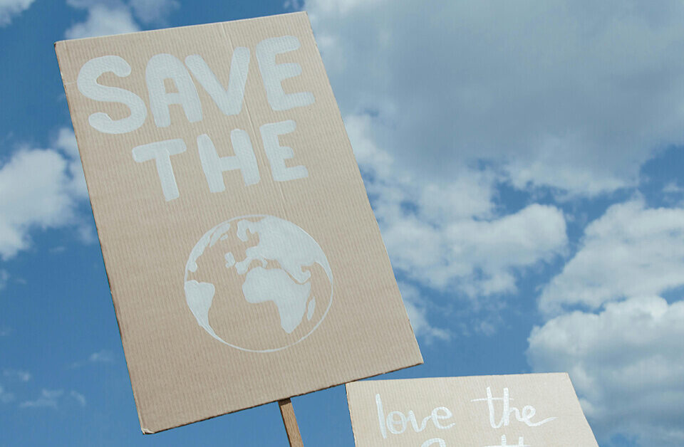 A person holding a cardboard sign that says "SAVE THE EARTH" with an illustration of the globe against a blue sky, representing the environmental goals of sustainable cannabis packaging.