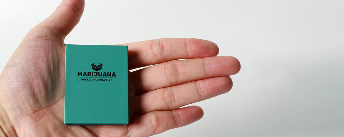 A close-up photograph of a small, custom-engineered teal paper carton held within a human hand, showcasing its compact size for discreet vape packaging solutions.