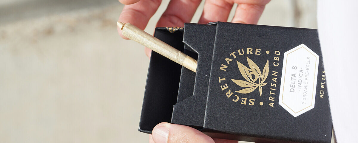A person's hands opening a rigid black cigar box containing a pre-roll, demonstrating premium custom marijuana packaging with gold foil branding for Secret Nature CBD.
