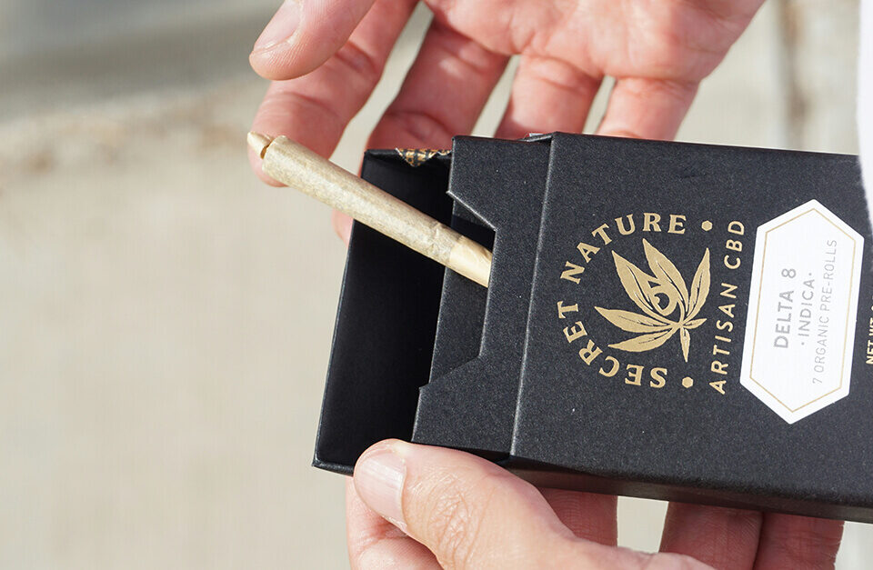 A person's hands opening a rigid black cigar box containing a pre-roll, demonstrating premium custom marijuana packaging with gold foil branding for Secret Nature CBD.