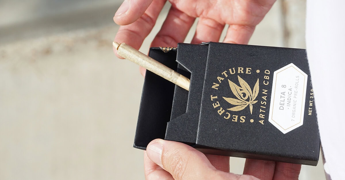 A person's hands opening a rigid black cigar box containing a pre-roll, demonstrating premium custom marijuana packaging with gold foil branding for Secret Nature CBD.