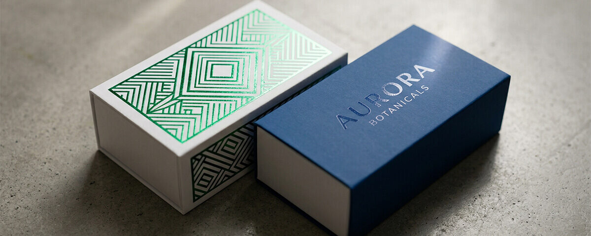 A detailed, close-up shot focusing on advanced finishing techniques used on custom sliding boxes. On the left, a white paperboard sleeve features intricate, reflective metallic green foil geometric patterns. On the right, a deep blue sleeve displays a subtle, glossy spot UV coating over the logo "AURORA BOTANICALS." Angled studio lighting highlights the varying textures and reflective properties of the packaging.