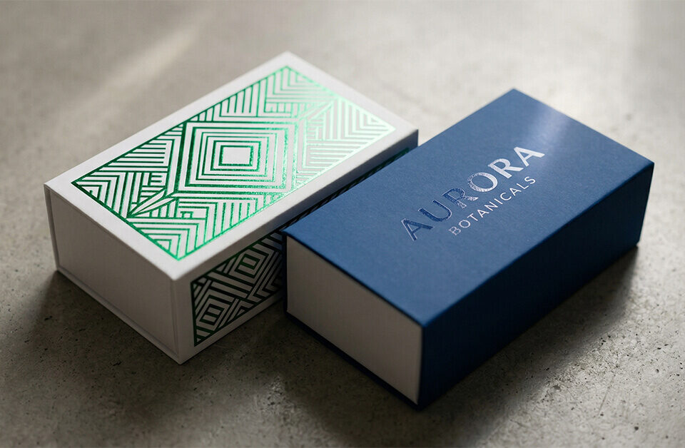 A detailed, close-up shot focusing on advanced finishing techniques used on custom sliding boxes. On the left, a white paperboard sleeve features intricate, reflective metallic green foil geometric patterns. On the right, a deep blue sleeve displays a subtle, glossy spot UV coating over the logo "AURORA BOTANICALS." Angled studio lighting highlights the varying textures and reflective properties of the packaging.