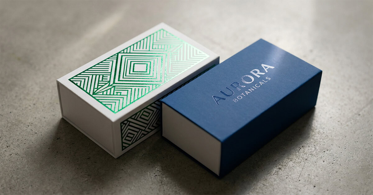 A detailed, close-up shot focusing on advanced finishing techniques used on custom sliding boxes. On the left, a white paperboard sleeve features intricate, reflective metallic green foil geometric patterns. On the right, a deep blue sleeve displays a subtle, glossy spot UV coating over the logo "AURORA BOTANICALS." Angled studio lighting highlights the varying textures and reflective properties of the packaging.