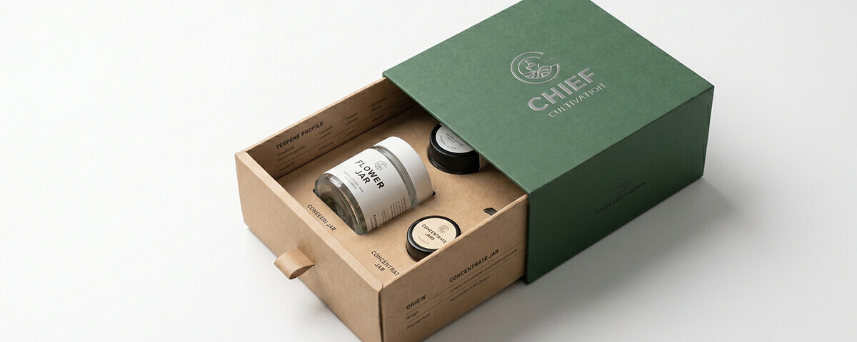 A partially open custom paper drawer box featuring a green outer sleeve and a kraft paper inner tray with custom inserts holding small glass jars, set against a solid white background.